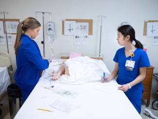 Two nurses standing over a mannequin practicing pokes.