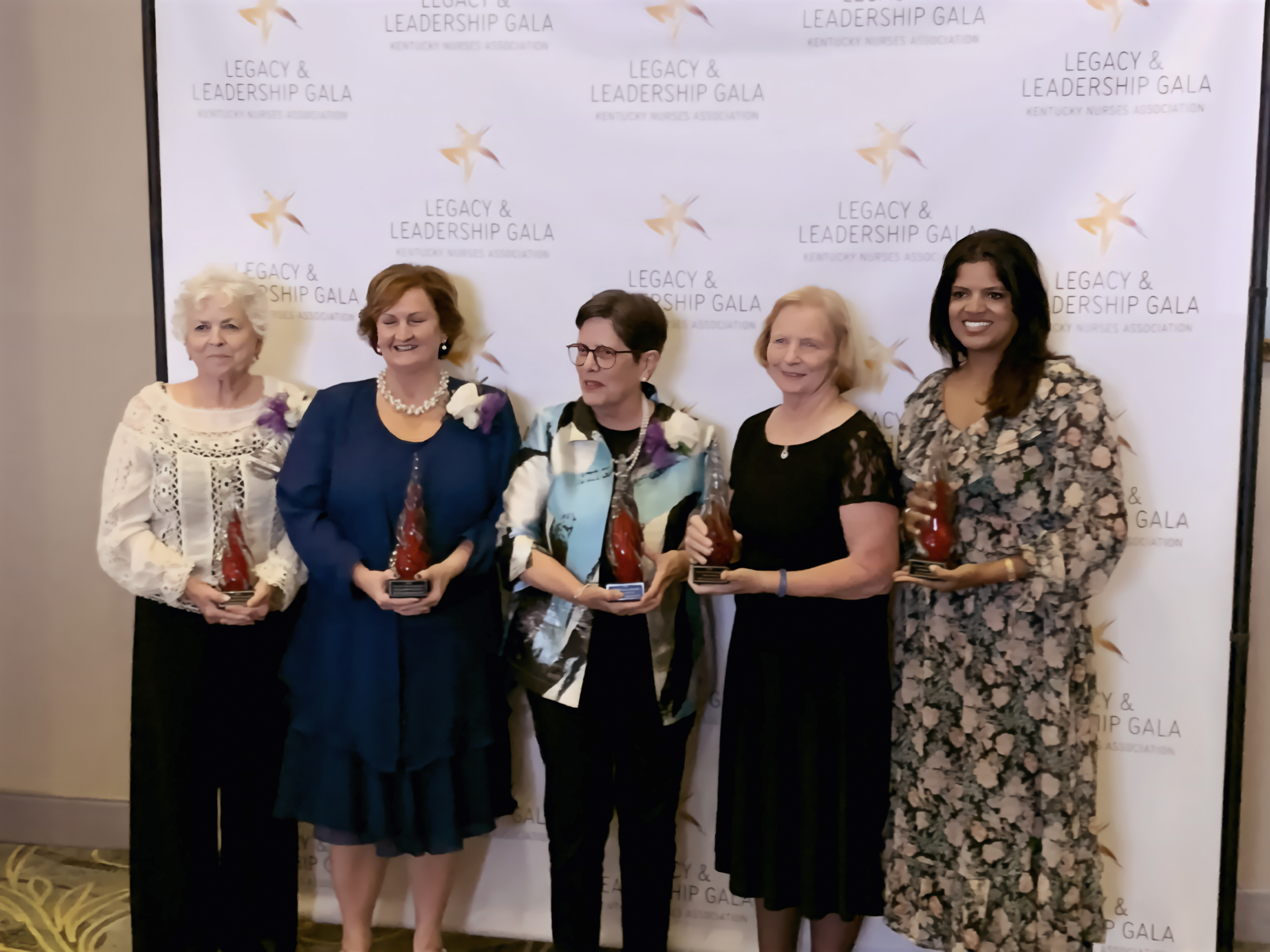 5 women standing together each holding a red glass trophy  