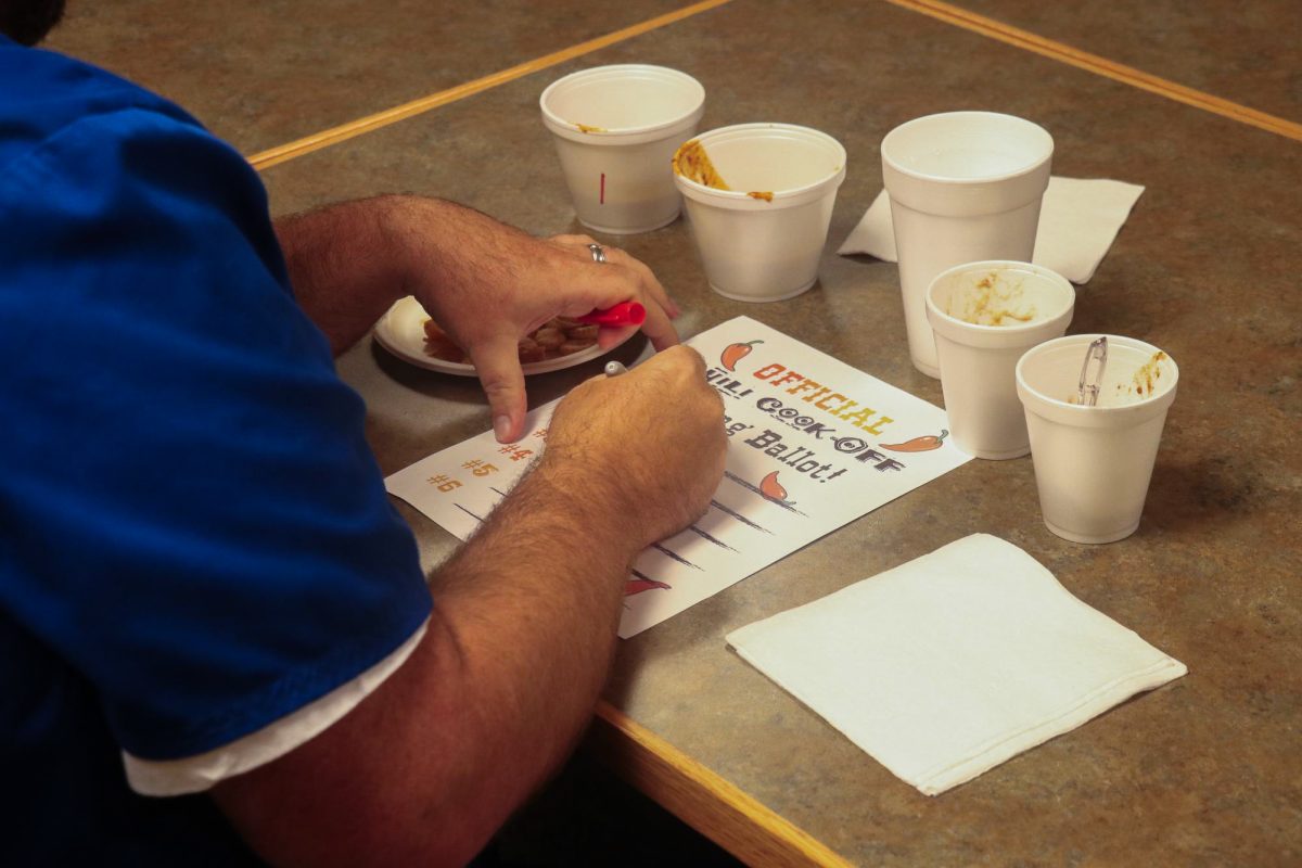 A photo of someone's hands writing on a piece of paper that reads "Official Chili Cookoff Voting Ballot." There are cups of chili on the table around the paper.