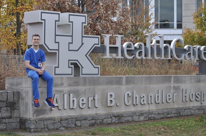 A man in nursing scrubs sits in front of a sign that says " UK HealthCare Albert B. Chandler Hospital."