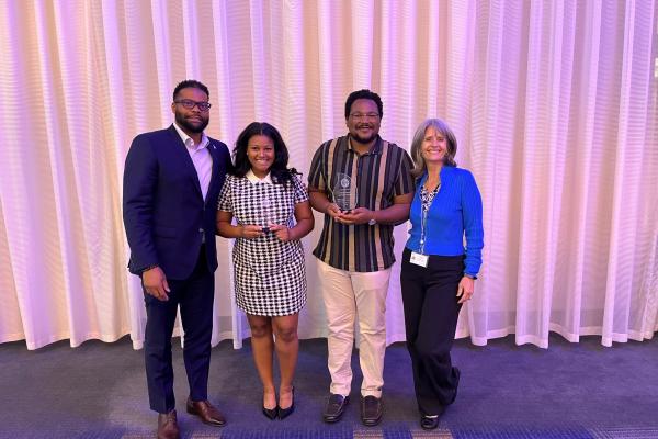 Photo of Corey Moore, Jordan Brown, Omar Brown, and Lee Anne Walmsley smiling. Jordan and Omar are holding awards. 