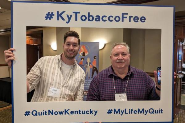 Two men smiling in a frame cutout that says "#KyTobaccoFree #QuitNowKentucky #MyLifeMyQuit"