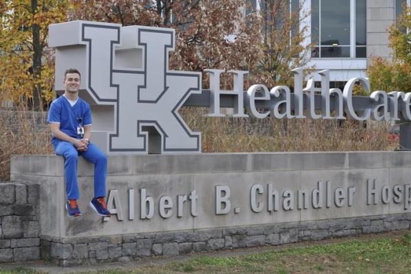 A man in nursing scrubs sits in front of a sign that says " UK HealthCare Albert B. Chandler Hospital."