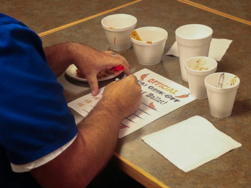 A photo of someone's hands writing on a piece of paper that reads "Official Chili Cookoff Voting Ballot." There are cups of chili on the table around the paper.