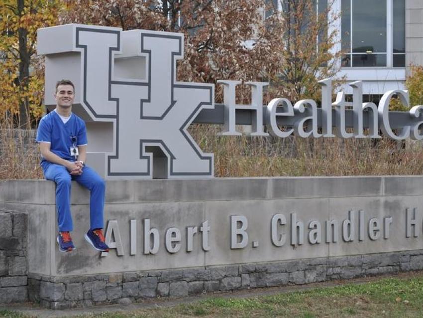 A man in nursing scrubs sits in front of a sign that says " UK HealthCare Albert B. Chandler Hospital."