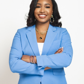 A Woman with curled black hair stands proudly in a light blue blazer with white shirt