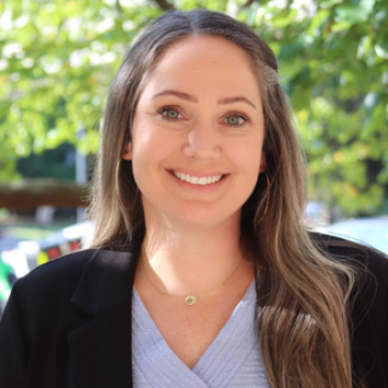 Woman smiling with long brown hair wearing a black blazer and blue shirt smiling on a bright day in front of green trees