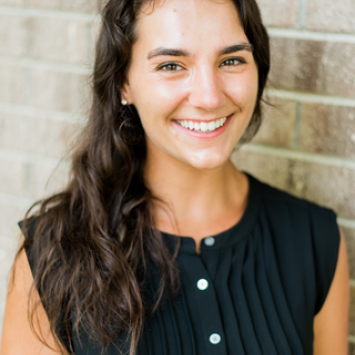 A woman with long dark curly hair wearing a black shirt stands, smiling in front of a brick wall