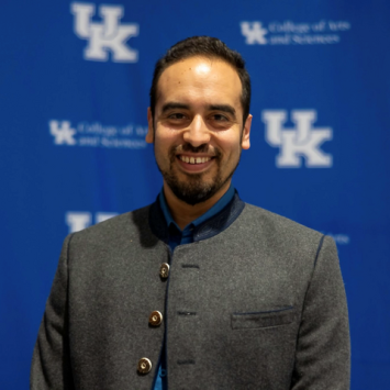 Man with black hair and a grey coat standing in front of a blue and white UK patterned background