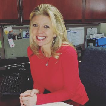 Woman with shoulder length blonde hair in a long sleeve red shirt sitting in an office with cabinets on the wall. She has her arms laid on a desk in front of her. She is smiling 