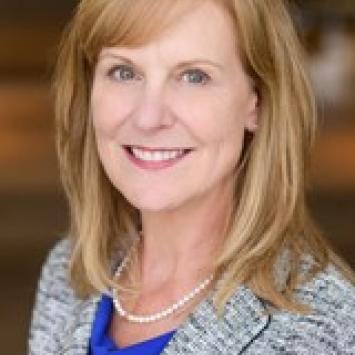professional headshot photo of a woman with reddish blonde hair that is shoulder length with short side bangs. She is wearing a grey and black heathered blazer with a royal blue undershirt and a pearl necklace. She is smiling, the background is blurred.