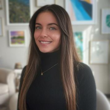 A photo of Victoria Thompson smiling and wearing a black sweater. Background shows pictures hanging on the wall. 