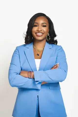 A Woman with curled black hair stands proudly in a light blue blazer with white shirt