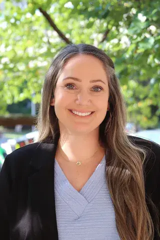 Woman smiling with long brown hair wearing a black blazer and blue shirt smiling on a bright day in front of green trees