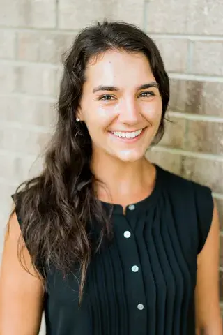 A woman with long dark curly hair wearing a black shirt stands, smiling in front of a brick wall