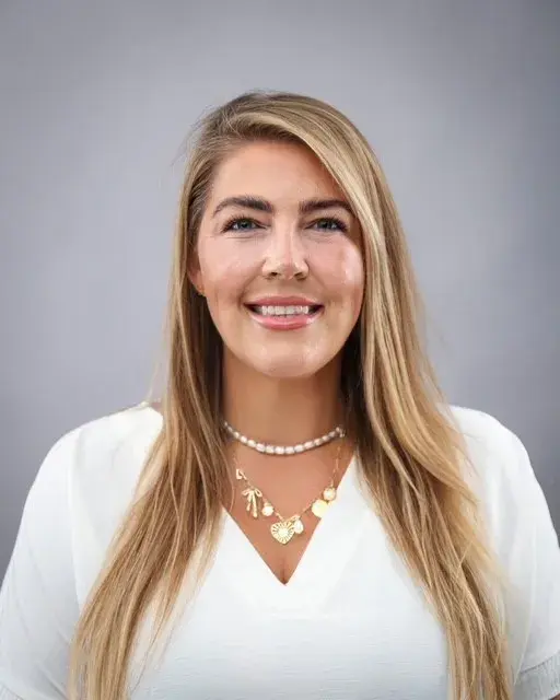 woman with long sandy blonde hair wearing a white shirt in front of a light grey background smiling 