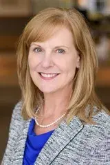professional headshot photo of a woman with reddish blonde hair that is shoulder length with short side bangs. She is wearing a grey and black heathered blazer with a royal blue undershirt and a pearl necklace. She is smiling, the background is blurred.