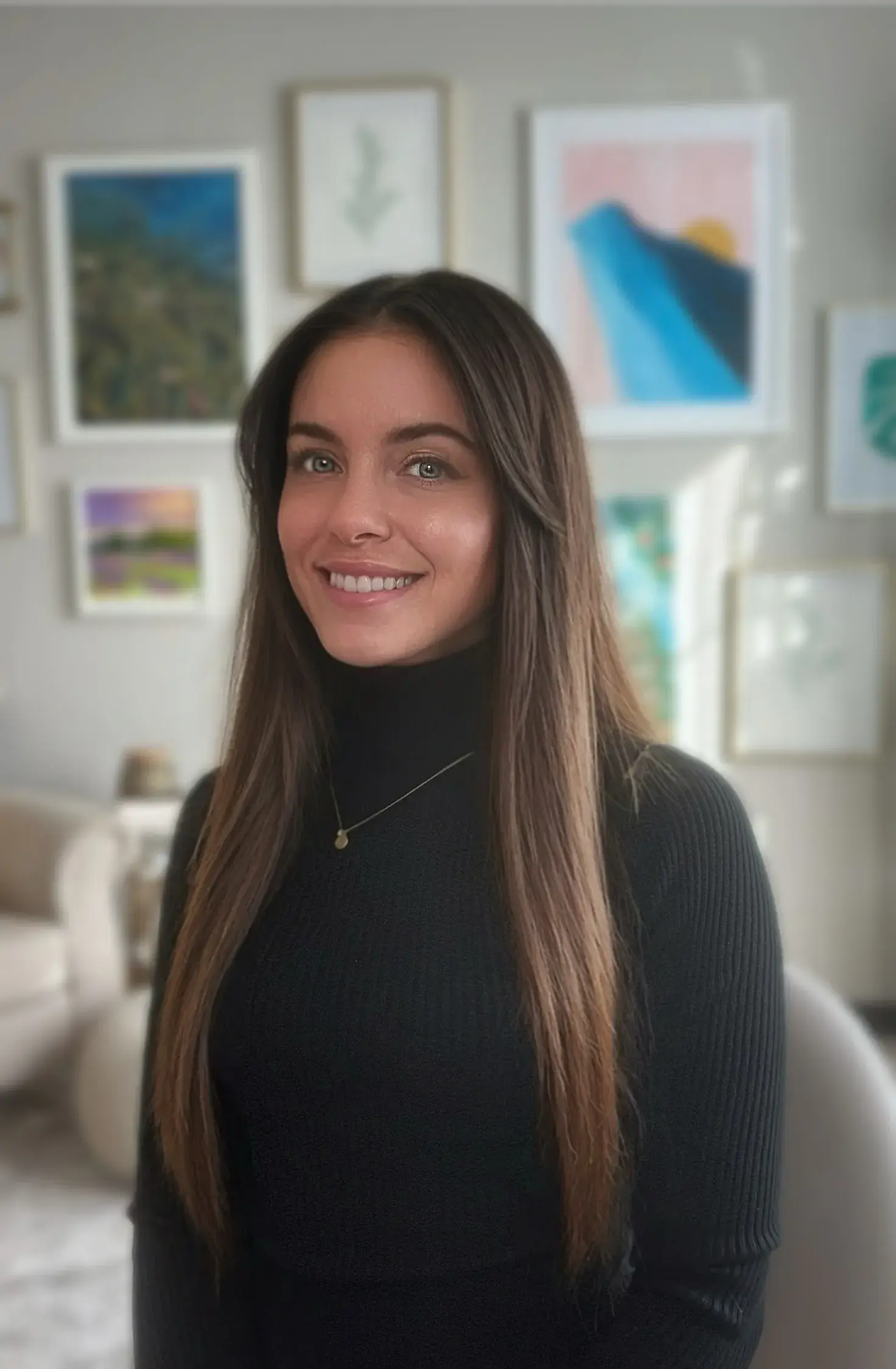 A photo of Victoria Thompson smiling and wearing a black sweater. Background shows pictures hanging on the wall. 