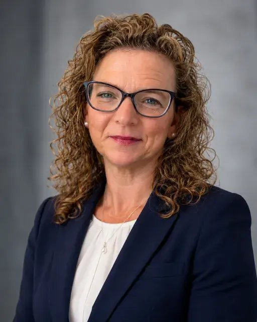 A headshot photo of a woman wearing glasses and a navy blazer, smiling. 